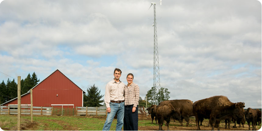 Small wind turbine on a buffalo ranch with couple standing in front of turbine and red barn.