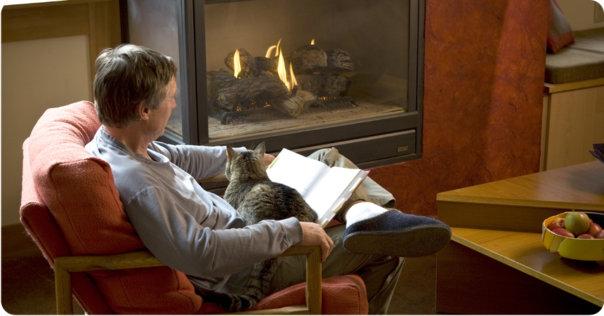 a man reading a book with a cat on his lap in front of a fireplace