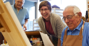 a laughing woman watching an elderly man painting