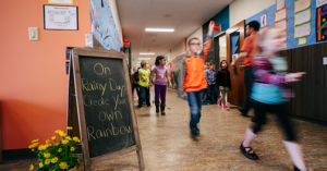 Kids walking down a hallway in a school with a chalkboard sign that says, "On Rainy Days Create Your Own Rainbow"