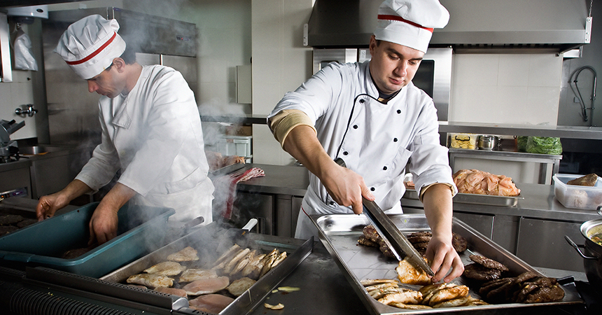 two chefs in a restaurant kitchen cooking chicken on a grill