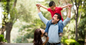 A latin man standing next to wife, carrying his son on his shoulders and smiling at each other in a horizontal medium shot outdoors.