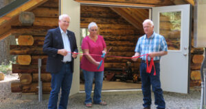 two men holding a ribbon and a woman with large scissors cutting it.