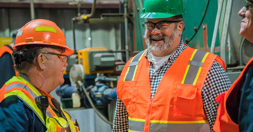 three men in bright orange vests and construction hats conversing and smiling