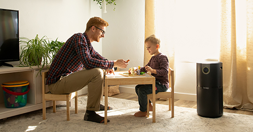 Man and child sit at small table playing with toys while an air purifier works nearby.