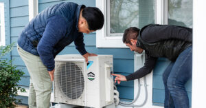 Two individuals inspecting an outdoor air conditioning unit mounted on the side of a blue house with white trim. One person is bent over examining the unit closely, while the other points at a component. Plants are visible in the foreground.
