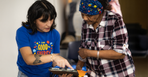 Two people engage with a tablet. The person on the left, wearing a blue T-shirt with the phrase “We have GOOD ENERGY,” points at the tablet screen, guiding the person beside them. The other person wears a plaid shirt and a vibrant headscarf, listening attentively. The setting suggests collaboration or instruction, emphasizing digital interaction and shared focus.