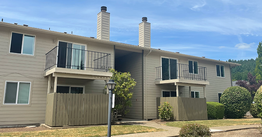 Two-story residential building with beige siding and two chimneys. Each unit features an upper-floor balcony and a fenced patio on the ground level. Multiple windows are visible on both floors. The building is surrounded by a neatly trimmed lawn and bushes, with a lamppost in the foreground. The sky is mostly clear with a few scattered clouds.