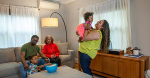A family enjoys time together in a living room. A man and an older woman sit on a couch with a young boy eating popcorn, while a woman in a yellow top lifts a smiling toddler in a pink dress.