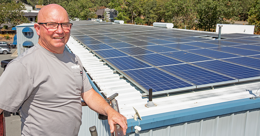 A man wearing glasses and a gray shirt stands smiling beside a building with a large array of solar panels covering most of its roof. The solar panels are arranged in neat rows. Trees and other buildings are visible in the background, suggesting a suburban or semi-urban setting. The image emphasizes renewable energy and sustainable development.