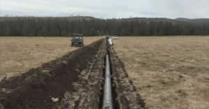 A wide trench cuts through a grassy field, with a large black pipeline being installed inside. Utility vehicles and a white truck are parked nearby, indicating active construction. In the background, coniferous trees and rolling hills suggest a remote, forested location.