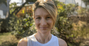 A person stands outdoors in bright sunlight, wearing a white sleeveless top. Their light-colored hair is styled back, and their face is warmly lit. Behind them, green trees and foliage frame a residential area with partially visible buildings, creating a serene and natural backdrop.