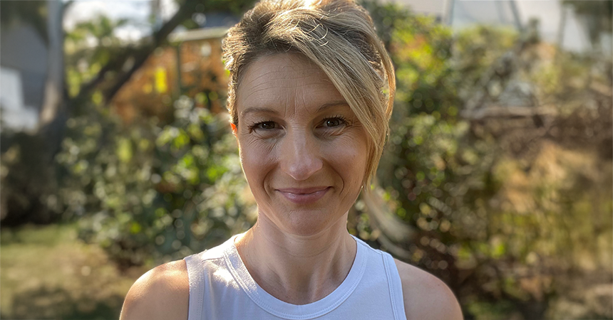 A person stands outdoors in bright sunlight, wearing a white sleeveless top. Their light-colored hair is styled back, and their face is warmly lit. Behind them, green trees and foliage frame a residential area with partially visible buildings, creating a serene and natural backdrop.