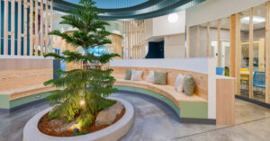Modern indoor space featuring a circular wooden seating area with neutral-toned cushions surrounding a planted tree. The background includes wooden slats, glass walls, and rooms with colorful chairs. The floor is polished concrete, and ceiling lights provide illumination.