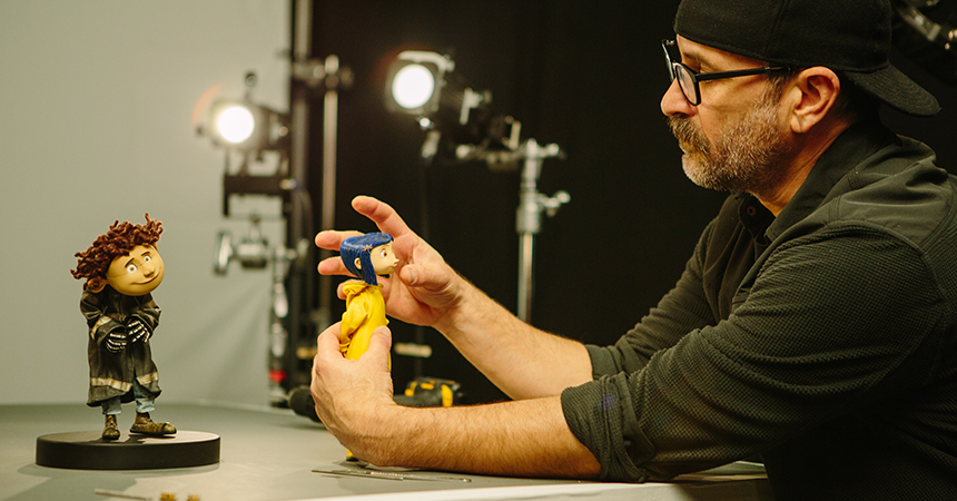 A man in a studio adjusts two detailed stop-motion puppets. He wears a backward black cap, glasses, and a dark long-sleeve shirt. One puppet resembles Coraline, dressed in a yellow raincoat and blue hat; the other has curly brown hair and a dark outfit, standing on a circular base. Studio lights and equipment are visible in the background.