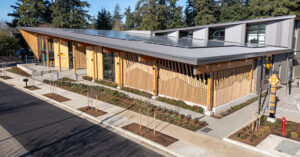 A modern eco-friendly building with a sloped roof covered in solar panels. The exterior features vertical wooden slats with geometric accents and large glass windows. Landscaped garden beds with young trees and shrubs line the paved sidewalk, and a small parking area is visible. Tall evergreen trees surround the site, emphasizing its natural setting.