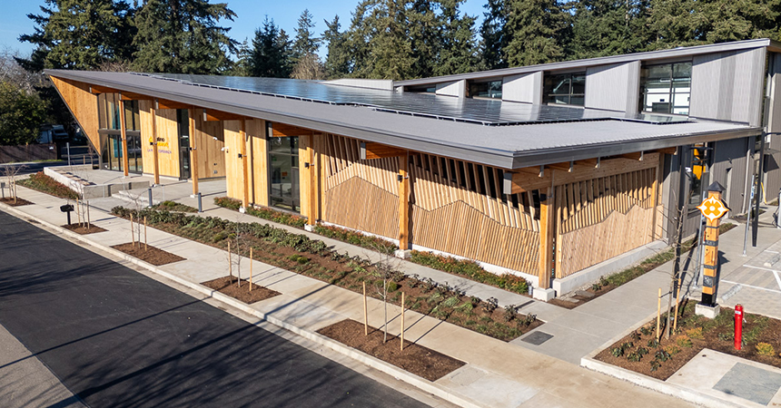 A modern eco-friendly building with a sloped roof covered in solar panels. The exterior features vertical wooden slats with geometric accents and large glass windows. Landscaped garden beds with young trees and shrubs line the paved sidewalk, and a small parking area is visible. Tall evergreen trees surround the site, emphasizing its natural setting.