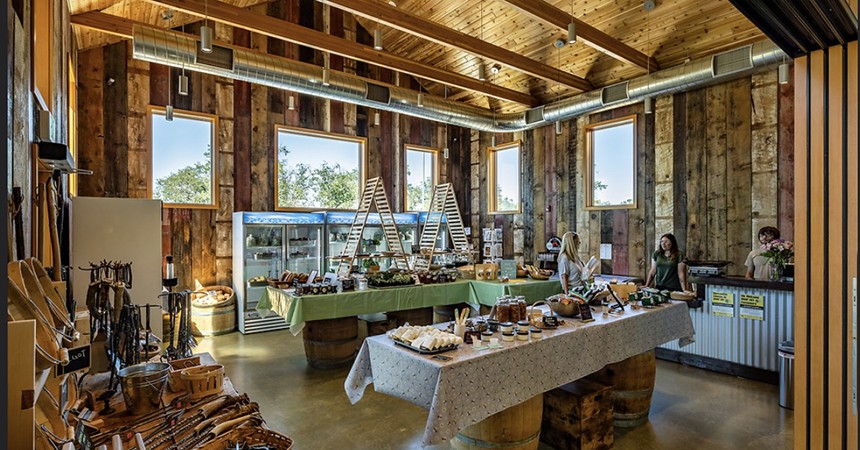 Interior of a rustic-style farm store with wooden walls, exposed beams, and large windows. Tables covered in green and white cloths display jars of preserves, baked goods, and artisanal items, supported by wooden barrels. Refrigerated cases and shelves line the back wall. Several people are present, including staff behind the counter and customers browsing. A section on the left features wooden utensils and tools. Natural light fills the space, highlighting the handmade, farm-to-table atmosphere.