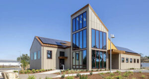 A modern municipal building labeled "Rusted City Hall" with beige walls, dark trim, and a tall glass-paneled section reflecting the surroundings. Solar panels line the roof, and young landscaping plants border a paved walkway leading to the entrance. The sky is clear and blue, suggesting a clean, well-maintained environment.