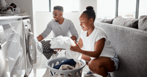Two people doing laundry together in a bright, modern living room. One person crouches in front of a front-loading washing machine, placing clothes inside. The other stands beside a laundry basket, holding a white garment. They are next to a gray couch, with large windows in the background letting in natural light. The scene conveys shared domestic responsibility and cooperation.