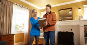 Two people stand in a warmly lit living room; one holds a clipboard and explains something to the other, who listens attentively. The room features a fireplace, mirror, large window with beige curtains, and decorative items.