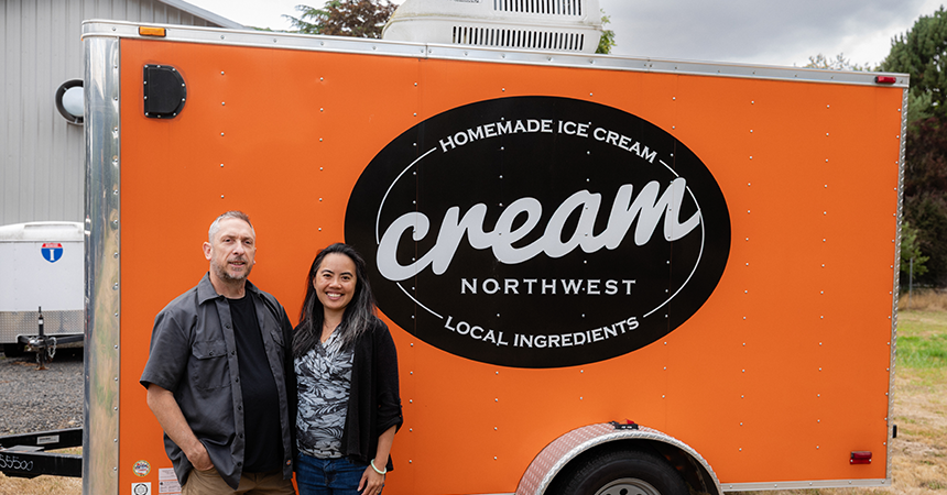 Two people stand in front of a bright orange food trailer with a large black oval sign that reads “Homemade Ice Cream,” “cream,” “Northwest,” and “Local Ingredients.” The word “cream” is stylized and centered, suggesting the business name “Cream Northwest.” The trailer promotes artisanal ice cream made with local ingredients.