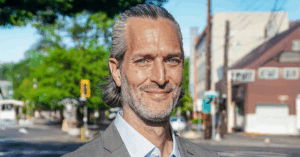 A middle-aged person with long, slicked-back gray hair and a beard stands outdoors on a sunny city street. They wear a gray suit jacket over a light blue dress shirt, gazing confidently ahead. Behind them, leafy green trees, a traffic light, and a mix of commercial and residential buildings line the urban backdrop. The lighting is bright and natural, suggesting midday.