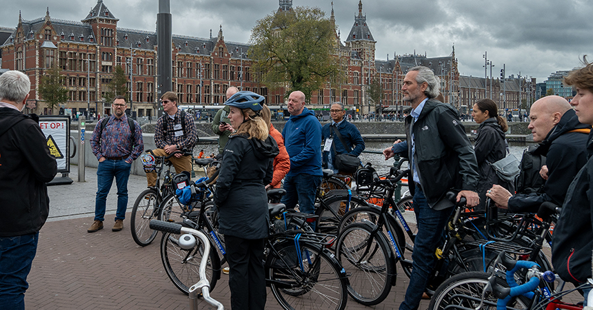 A group of cyclists gathers in an urban square near a historic brick building with spires and a clock tower, possibly a train station or government site. The participants, dressed in casual outdoor clothing and some wearing helmets, appear to be listening to a guide or organizer. Bicycles are parked or held nearby, suggesting a group ride or tour. The sky is overcast, and a Dutch-language warning sign ("LET OP!") is visible in the background, indicating the location may be in the Netherlands. The scene conveys communal activity, cultural tourism, and sustainable urban mobility.
