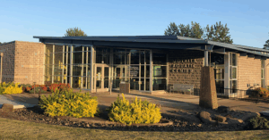 Exterior view of the Molalla Aquatic Center on a clear day, featuring a modern building with large glass windows, a stone facade, and a flat roof with extended eaves. The entrance is framed by vibrant landscaping with yellow and red flowers and a stone pathway, set against a backdrop of tall trees and blue sky.