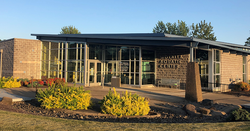 Exterior view of the Molalla Aquatic Center on a clear day, featuring a modern building with large glass windows, a stone facade, and a flat roof with extended eaves. The entrance is framed by vibrant landscaping with yellow and red flowers and a stone pathway, set against a backdrop of tall trees and blue sky.