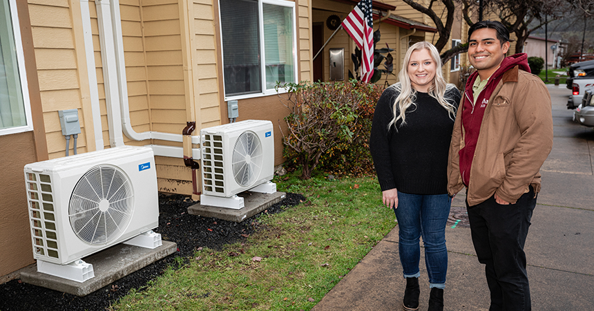 Two people stand outside a beige residential building near two Daikin-branded HVAC units mounted on concrete pads. The building has white-framed windows, an American flag by the entrance, and neatly trimmed shrubs along a sidewalk in the foreground. The scene suggests a home energy upgrade or heat pump installation.
