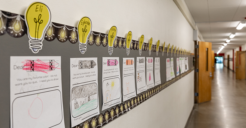 School hallway display featuring student artwork and writing assignments mounted on a wall. Each piece includes a yellow paper light bulb with a student's name and a letter addressed to a favorite color, expressing affection and a plea for the color not to disappear. The display is bordered with a decorative light bulb trim, emphasizing creativity and bright ideas.