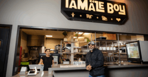 Interior of Tamale Boy restaurant with an open kitchen and stainless steel shelves. A staff member stands behind the counter, facing a person in front. Above them, a bold illuminated sign reads “TAMALE BOY,” flanked by icons of an ear of corn, tamale, mask, and heart. The space is clean, modern, and welcoming.