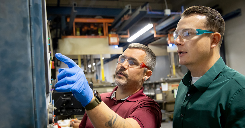 Two people wearing safety glasses stand in an industrial workshop. The person in a maroon shirt and blue gloves points at a machine control panel while explaining something to the person beside them, who wears a green shirt. Large equipment and overhead lighting fill the background, suggesting a manufacturing or technical training environment.