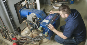 A technician kneels in front of a large blue industrial boiler control panel, adjusting switches and buttons. The panel is covered with gauges, indicator lights, and wiring, and is connected to surrounding pipes and valves in a mechanical room with concrete flooring and additional equipment in the background.