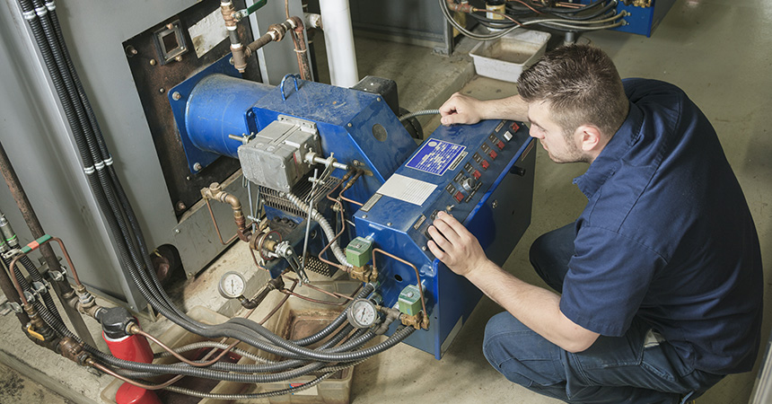 A technician kneels in front of a large blue industrial boiler control panel, adjusting switches and buttons. The panel is covered with gauges, indicator lights, and wiring, and is connected to surrounding pipes and valves in a mechanical room with concrete flooring and additional equipment in the background.