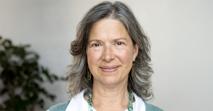 A person with shoulder?length gray hair smiles softly at the camera. They wear a white top, a green cardigan, and matching green beaded jewelry. A leafy plant is blurred in the background against a light-colored wall.
