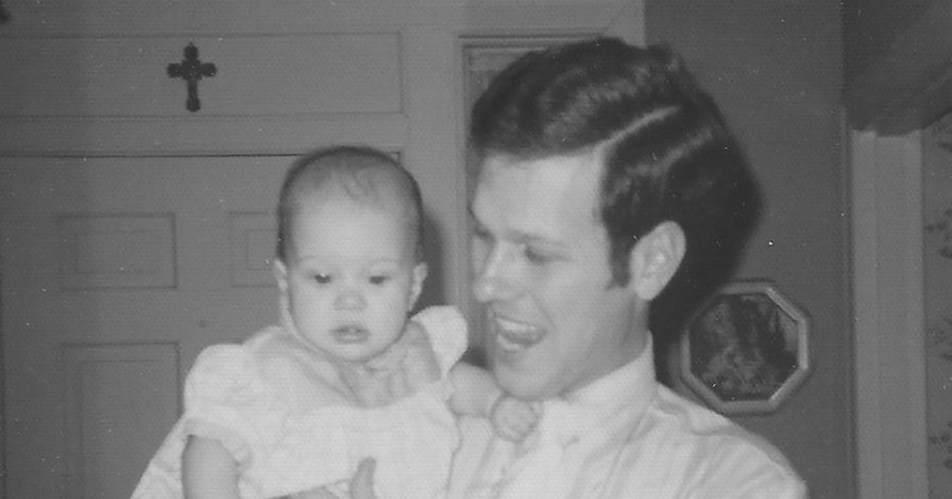 Black?and?white indoor photograph of an adult holding a baby. The adult, wearing a collared shirt and tie, looks down at the child with a gentle expression. The baby, dressed in a light outfit with puffed sleeves, faces the camera. Behind them is a door with a cross mounted above it and a framed picture on the wall.