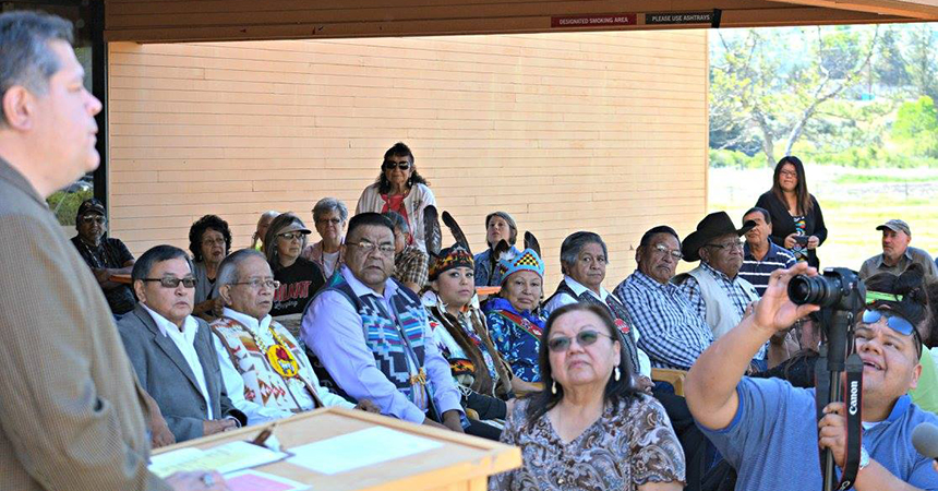 A man stands at a podium speaking to an audience gathered under an outdoor covered structure. The crowd includes people in both everyday clothing and traditional Native American regalia, such as beaded garments and feathered accessories. One person in the front records the event with a Canon camera. A sign overhead reads “Designated Smoking Area — Please Use Ashtrays.” The atmosphere suggests a cultural or community ceremony.