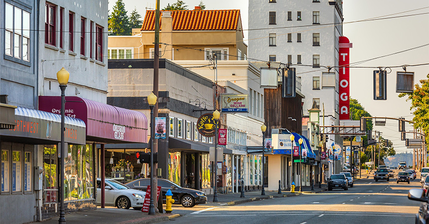 Downtown streetscape with storefronts, parked cars, and streetlights lining a wide road. A tall red vertical “TIOGA” theater sign stands above the buildings, while overhead utility lines and traffic signals cross the street. The scene is illuminated by warm late-afternoon sunlight with a few vehicles traveling in the distance.