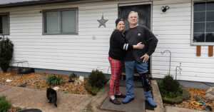 Two people stand on a concrete path in front of a white house with horizontal siding. One wears a black long?sleeve shirt and red plaid pants; the other wears a dark gray shirt, blue jeans, and a medical boot with a leg brace on the right leg. A black cat walks along the path to the left. Behind them are small landscaped garden beds with mulch, rocks, and shrubs, a black front door with a decorative star on the wall, and two windows.