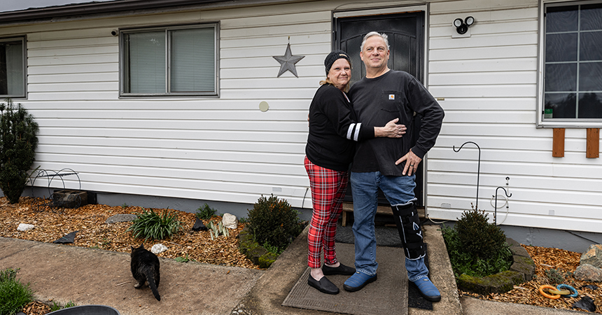 Two people stand on a concrete path in front of a white house with horizontal siding. One wears a black long?sleeve shirt and red plaid pants; the other wears a dark gray shirt, blue jeans, and a medical boot with a leg brace on the right leg. A black cat walks along the path to the left. Behind them are small landscaped garden beds with mulch, rocks, and shrubs, a black front door with a decorative star on the wall, and two windows.