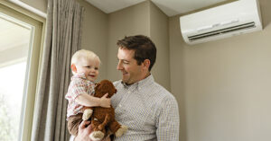 An adult wearing a plaid shirt holds a smiling young child who is also dressed in plaid and clutching a stuffed animal. They stand indoors near a window with gray curtains, and a white wall?mounted air?conditioning unit is visible behind them. The scene feels warm and domestic.