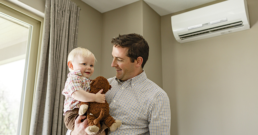 An adult wearing a plaid shirt holds a smiling young child who is also dressed in plaid and clutching a stuffed animal. They stand indoors near a window with gray curtains, and a white wall?mounted air?conditioning unit is visible behind them. The scene feels warm and domestic.