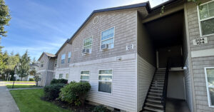 A two?story apartment building with light-colored siding and exterior stair access. The lower level has horizontal paneling and the upper level has shingle-style siding. A central staircase leads to the second-floor walkway. Signs on either side of the stairs display apartment numbers ‘203 103’ and ‘204 104.’ Several windows are visible, some with window air-conditioning units. The building is surrounded by a neatly maintained lawn, shrubs, and mature trees.