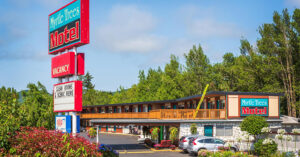 A two?story roadside motel called “Myrtle Trees Motel” sits among tall, leafy trees and landscaped shrubs. The building has a wooden balcony on the upper level and a stone façade on the lower level. A large sign out front reads “Myrtle Trees Motel,” with smaller signs for “No Vacancy,” “Clean Rooms & Scenic Views,” and “AAA Approved.” Several parked cars line the front of the building, and the sky is partly cloudy with patches of blue.