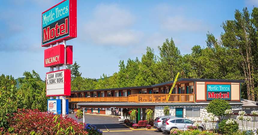 A two?story roadside motel called “Myrtle Trees Motel” sits among tall, leafy trees and landscaped shrubs. The building has a wooden balcony on the upper level and a stone façade on the lower level. A large sign out front reads “Myrtle Trees Motel,” with smaller signs for “No Vacancy,” “Clean Rooms & Scenic Views,” and “AAA Approved.” Several parked cars line the front of the building, and the sky is partly cloudy with patches of blue.