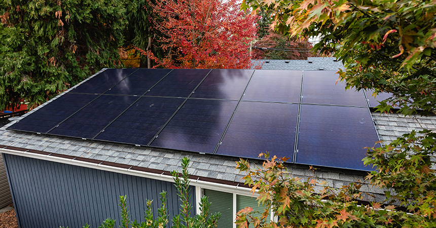A residential house with a shingled roof fitted with twelve solar panels arranged in three rows of four. The home is surrounded by trees with orange and yellow autumn foliage, and the panels are angled to capture sunlight.