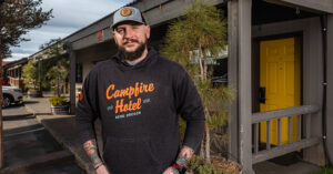 A person in a dark hoodie reading “Campfire Hotel – Bend, Oregon” stands outside a building with a yellow door and wooden railing. They wear a gray cap and have tattooed arms, with a parking area and trees behind them.