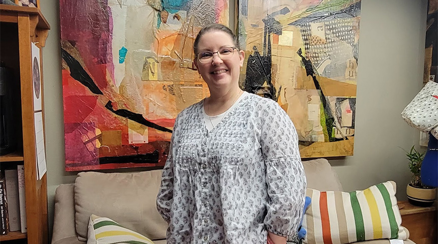 A person stands indoors in front of a beige couch with striped pillows. Behind them hangs a large abstract painting with bold red, orange, black, and blue shapes. A wooden shelf with books is on the left, and a lamp with a patterned shade and blue base is on the right.